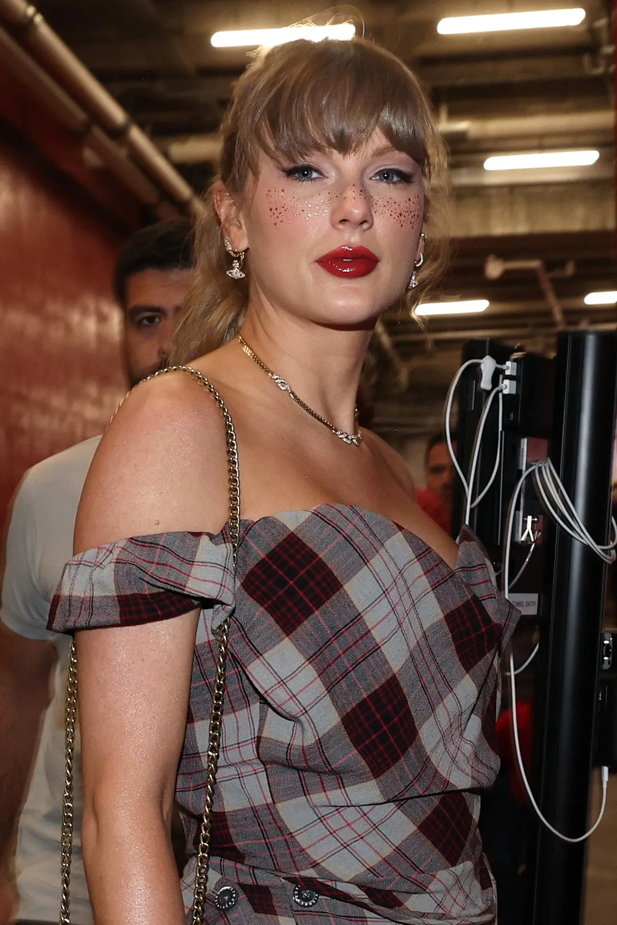 Taylor Swift na futbalovom zápase v kockových šatách s červeným rúžom a trblietavými pehami.
